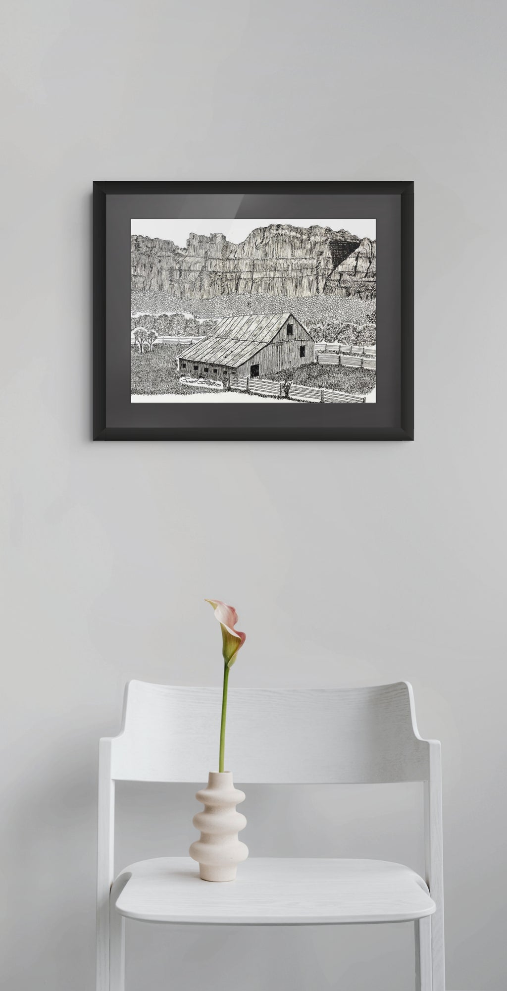A framed black and white landscape art print of a rustic cabin in a canyon, displayed on a light gray wall above a white chair with a small vase holding a single pink calla lily.