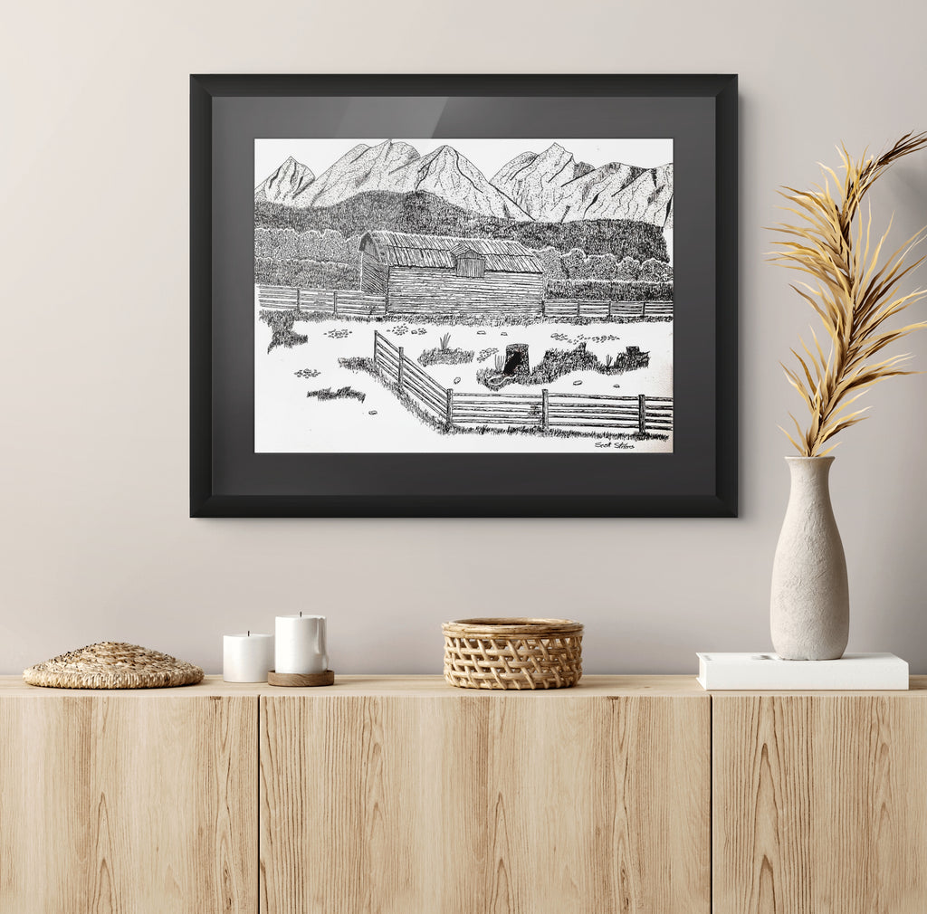A framed black and white landscape art print depicting a rustic farm scene with mountains, hung on a light beige wall above a wooden console table styled with candles, a basket, and a vase of dried palm leaves.