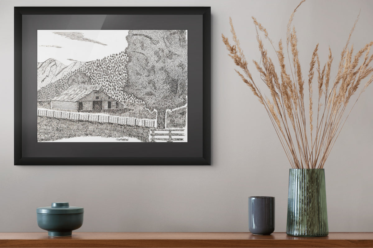 A framed black and white landscape art print hangs on a light gray wall above a wooden shelf. The print depicts a small cabin, fence, and mountains.